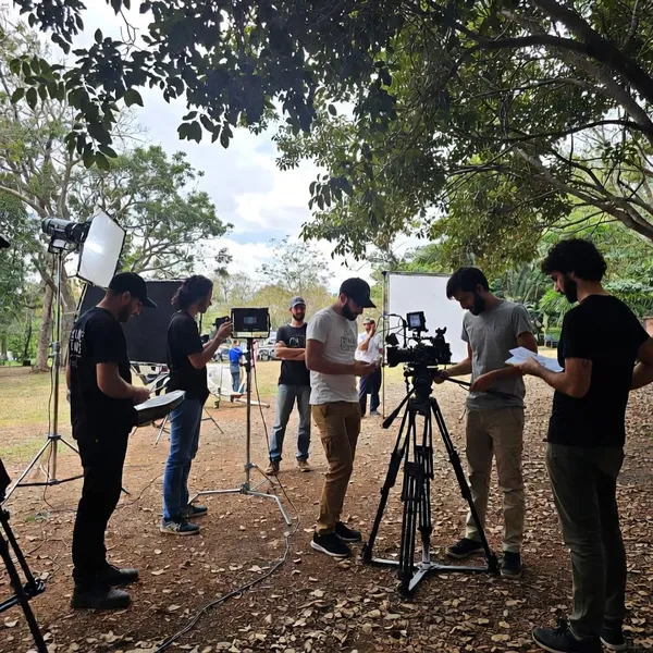 Film crew setting up a camera and lights outdoors under trees